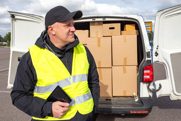 Delivery worker in a high-visibility vest stands beside an open van loaded with cardboard boxes, holding a tablet and preparing for the day’s deliveries.