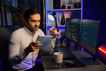 Smart IT developer working on software development coding on pc screen and reading note of newest application project, thinking program update online website data at neon light cyber office. Surmise.