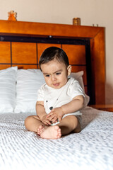 Baby sitting on bed with an angry face trying to reach his feet and wearing white clothes