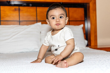 Baby sitting in bed, turning to one side, blowing a kiss, and wearing white clothes