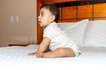 Baby sitting on a bed, leaning his arms on the bed, looking to the side with a surprised face
