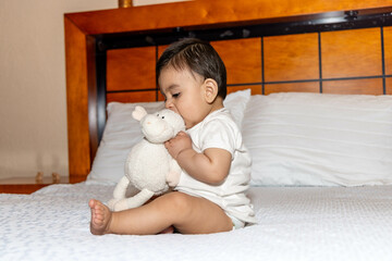 Baby sitting on a bed with a stuffed animal, baby biting a stuffed animal on a bed