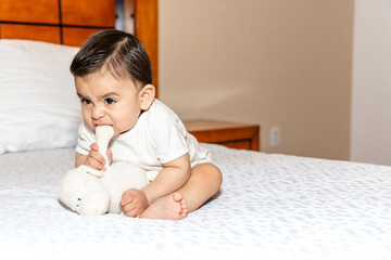 Baby sitting and half leaning on a bed, biting a stuffed animal to vent his anger, the baby makes angry gestures