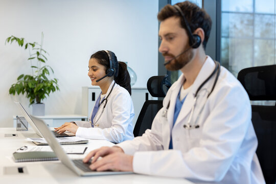 Two healthcare professionals wearing headsets and lab coats, working on laptops in a modern office setting. They are smiling while using technology.