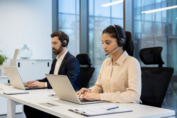 Two call center employees, a woman and a man, are working on laptops at their desks while wearing headsets.