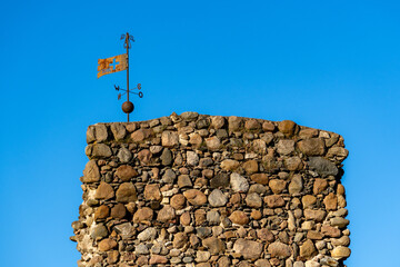 Old stone wall with weathered metal weather vane against clear blue sky. Traditional rural...