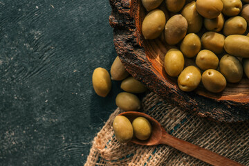 Rustic wooden bowl filled with green olives on burlap fabric with olive branch in background....