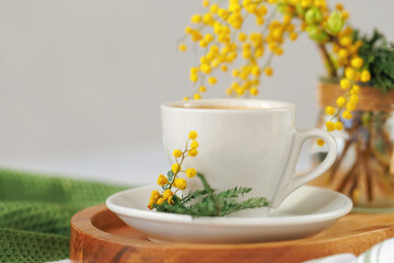 A cup of coffee sits on a wooden tray, accompanied by fresh mimosa flowers, creating a cozy and elegant spring ambiance.
