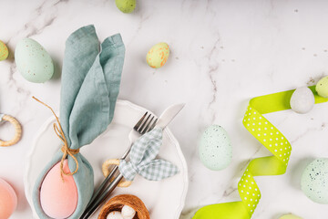 Top view of festive plate setting with green napkin bunny, silver cutlery, speckled eggs and green ribbon on white marble surface