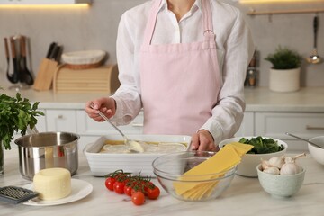 Woman spreading bechamel sauce onto spinach lasagna at marble table in kitchen, closeup