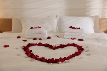 Heart made with rose petals and burning candles on bed indoors