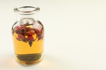 Open perfume bottle with aromatic essential oil and dried roses on white table, closeup. Space for text