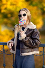 Happy pretty girl with lollipop in hand and sunglasses. Young woman with colorful candy at sunset in leather jacket