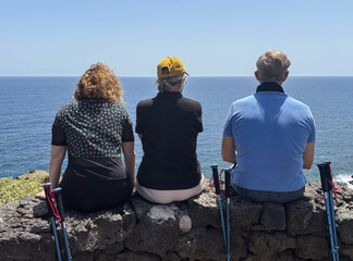 Fototapeta premium Rear view of three bonding friends sitting face the sea in a trekking sunny day admiring the horizon over water