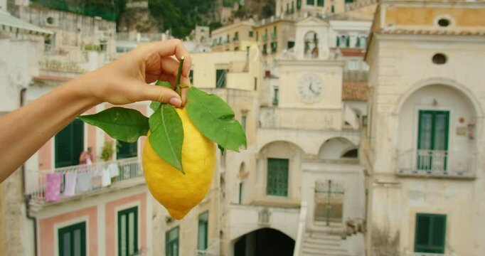 Large Sfusato Amalfitano lemon held in hand above pastel buildings in Atrani city on Amalfi coast on a summer morning