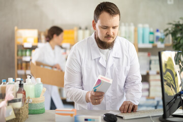 Adult male pharmacist holds a package of pills in hands and stands behind the counter near the computer. Pharmacist works in a pharmacy against the background of an assistant who puts goods on the