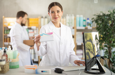 Young woman pharmacist with package of pills, she stands behind the counter near the computer. Pharmacist works in a pharmacy against the background of an male assistant who puts goods on the shelves