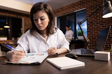 Fototapeta premium Focused female executive checking business reports on a clipboard at her desk. Dedicated businesswoman holding a pen and reviewing financial documents in a brick wall office.