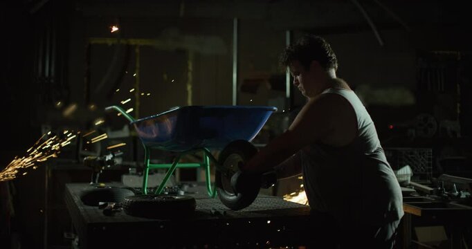 Intense looking man working on a wheel barrow in metal shop with sparks flying around him - dolly in