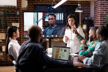 Multiethnic startup team in modern office with brick wall background, engaged in late night business meeting. A digital screen displays video call with remote african american manager.