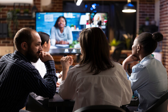 Business professionals collaborate in nighttime office meeting. A videoconference call is displayed on digital monitor as team members discuss marketing strategies and review financial data.