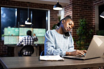 Black female employee manages multiple tasks at once, talking on her cellphone while typing financial data on laptop. African american woman using her personal computer and mobile device in office.