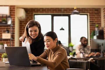Multiethnic employees working together at office desk, reviewing financial data on laptop. An inclusive workspace where caucasian manager and asian wheelchair user discuss business growth strategies.