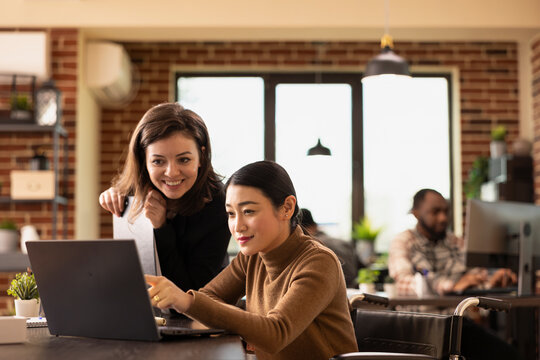 Multiethnic employees working together at office desk, reviewing financial data on laptop. An inclusive workspace where caucasian manager and asian wheelchair user discuss business growth strategies.