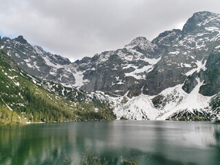 Naklejka premium Snow-covered Tatra Mountains surround the emerald waters of Morskie Oko lake, with dense pine forests and dramatic rocky slopes under cloudy skies