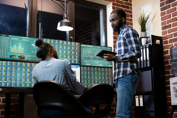 African American forex traders exchange investment strategies while tracking real time stock market transactions during night shift in brick wall office. Teamwork discussion at tech savvy workstation.