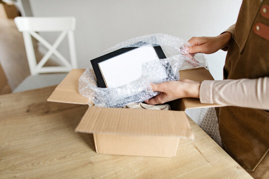 Close up of woman small business owner wrapping photo frame, packing box with fragile product into protective tape prepare parcel for shipment delivery