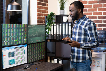 African american male investor standing with his tablet, monitoring forex charts on multiple screens. The office features a brick wall, creating a financial tech savvy workspace.