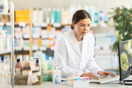 Qualified female pharmacist works at a computer in a European pharmacy. Pharmacist checks the availability of goods in a spreadsheet against the background of shelves with medicines