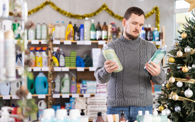 In Christmas decorated shop, man examines product and doubts, chooses dishwashing soap, study items properties in household department. Buyer examines and selects product..