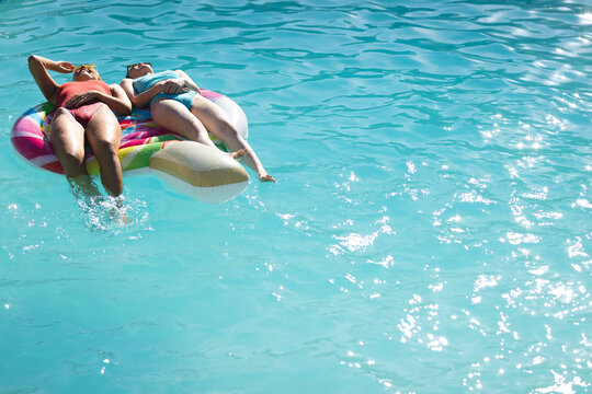 Diverse friends lounging on colorful ice cream cone float in outdoor blue pool under bright sun, cop