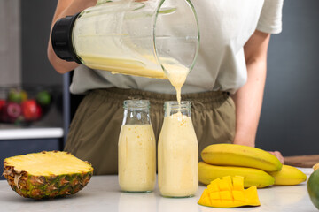 Woman pouring refreshing tropical smoothie with banana, pineapple and mango from blender into bottle