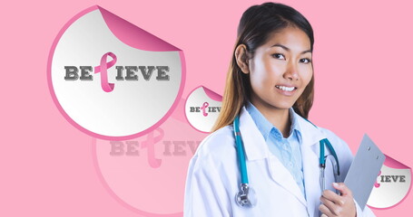 Image of believe text with pink ribbon over smiling biracial female doctor on pink background