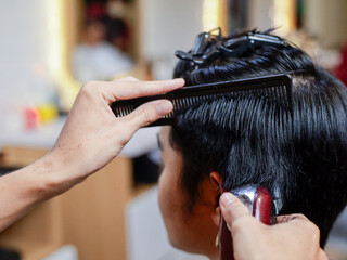 Fototapeta premium Close-up of Asian man getting a haircut at a barbershop. A barber uses a red electric clipper and black comb to trim the side hair. Background shows blurred salon environment and warm tones