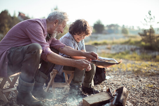 Grandfather and grandson preparing fish while camping by river in forest