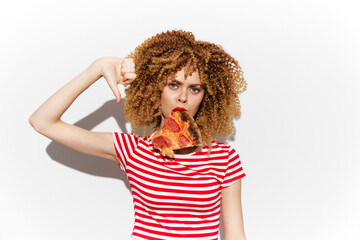 Young woman with curly hair making a disapproving gesture while holding a slice of pizza, wearing a...