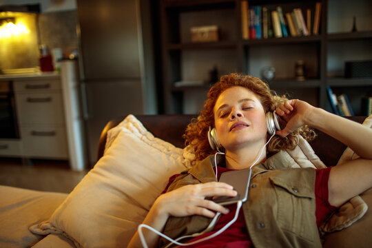 Young woman relaxing on couch with headphones and smartphone at home - Powered by Adobe