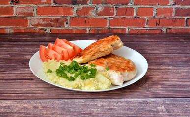 Grilled chicken breast fillet with mashed potatoes and tomatoes on a wooden table.