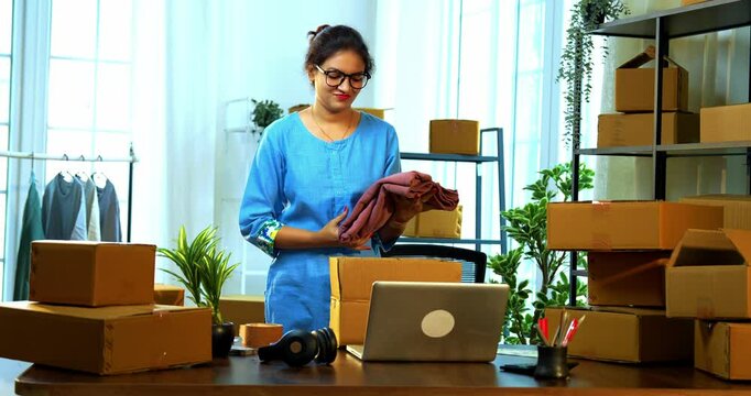 Young Indian Asian small businesswoman in store room office packing garments into cardboard carton box on desk for shipment, preparing online export dispatch order for delivery logistics and shipping