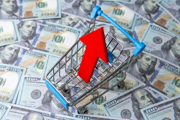 A shopping cart and red upward pointing arrow on a bed of blurred one hundred U.S. dollar bills....