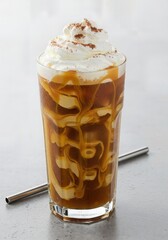 This is a vertical view of a tall glass of iced coffee with creamy swirls of caramel drizzle, topped with whipped cream and a dusting of cocoa powder, served with a metal straw.