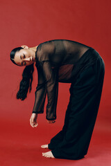 Fashion asian woman in sheer top and black pants bending over on red background with hands on knees...