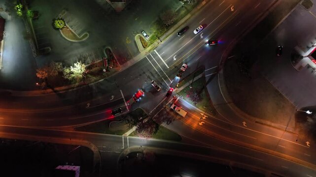Aerial view of a busy intersection at night, illuminated by the lights of emergency vehicles. Traffic is halted as responders manage scene. Nearby, empty parking lot contrasts with the active roadway.