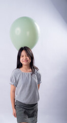 Asian girl with long black hair smiles with eyes closed while standing indoors under a large green balloon floating above her head, wearing a striped blouse and dark skirt against white background