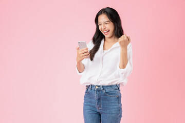 Cheerful young Asian woman holding smartphone with fists clenched celebrating victory expressing...