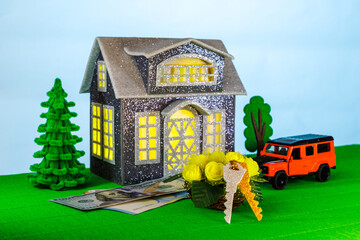 On a light background with yellow light in the windows, a gray house surrounded by an orange car and flowers with keys and euro and dollar banknotes